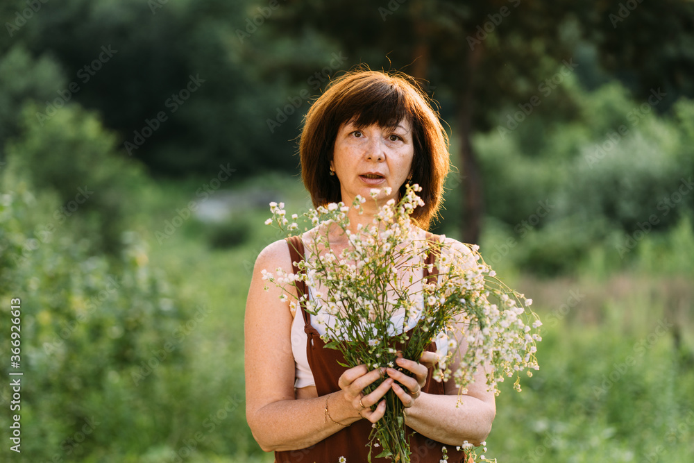 Portrait of middle age mature woman in brown overalls with a bouquet of chamomile herbs hot sunny summer, sunset in village. Life style without face concept. Menopause and changes in body. Portrait of middle age mature woman in brown overalls with a bouquet of chamomile herbs hot sunny summer, sunset in village. Life style without face concept. Menopause and changes in body.