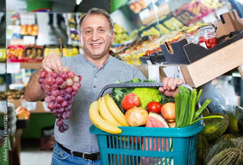 Smiling cheerful positive glad male is standing with basket with vegetables and fruits in the supermarket.