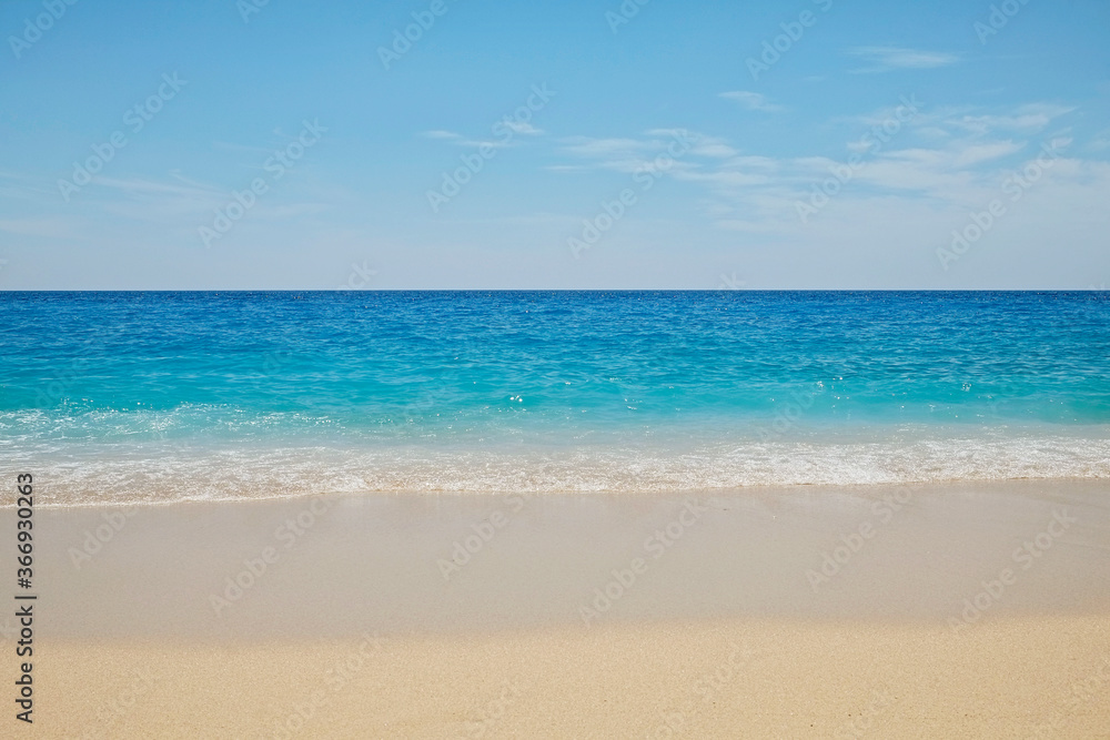 Soft ocean wave with foam licking the beautiful golden sandy beach shore. Close up of turquoise clear sea water texture on a windless and sunny summer day. Tropical background, copy space, top view.