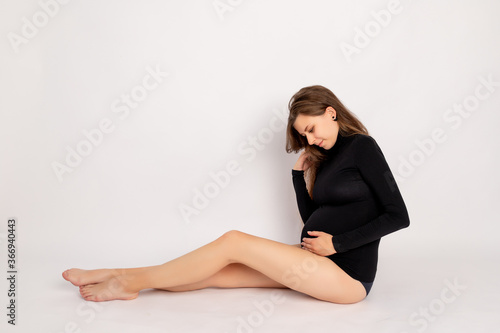 beautiful pregnant girl in a black bodysuit sitting on a white background holding her stomach, profile photo, fashion shoot of the future young mother, place for text