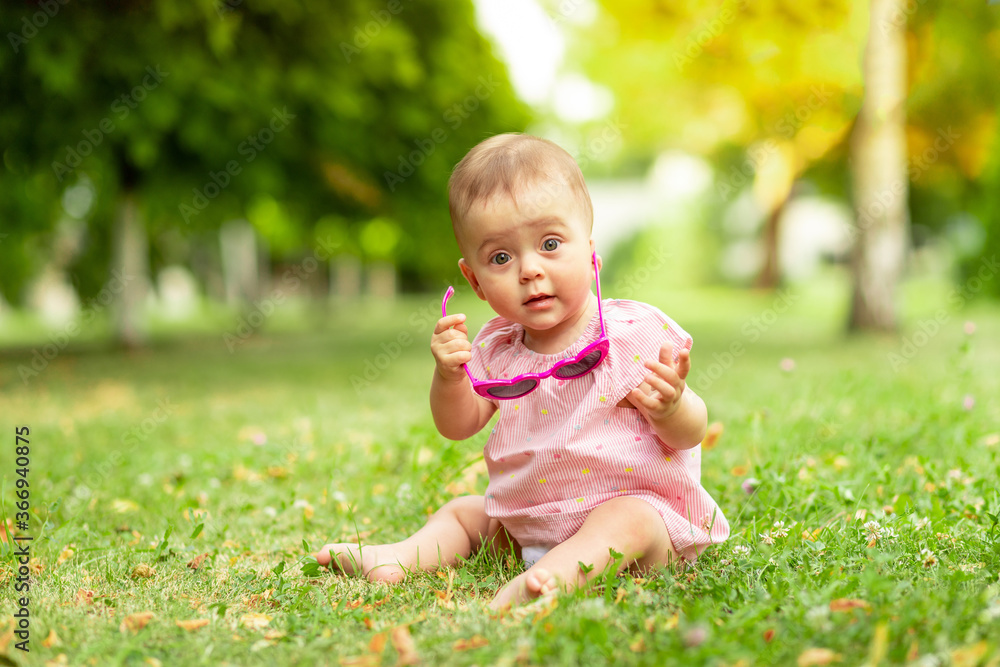 little baby girl 7 months old sitting on the green grass in a pink bodysuit and bright glasses, walking in the fresh air little baby girl 7 months old sitting on the green grass in a pink bodysuit and bright glasses, walking in the fresh air