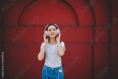 Young attractive hipster girl listening music in headphones while standing on red wall background,pretty female traveler in t-shirt with copy space for frand name enjoying favorite playlist outdoors photo