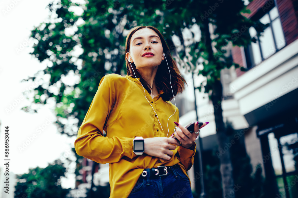 Half length portrait of confident student looking at camera while listening to favorite radio station via earphones and application on cellular.Cute hipster girl spending free time with gadget outside Half length portrait of confident student looking at camera while listening to favorite radio station via earphones and application on cellular.Cute hipster girl spending free time with gadget outside
