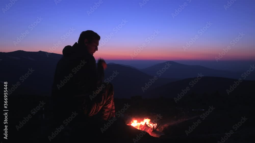 A man sits at night by a fire in the mountains. A tourist admires the landscape and looks at the burning fire. A man sits at night by a fire in the mountains. A tourist admires the landscape and looks at the burning fire.