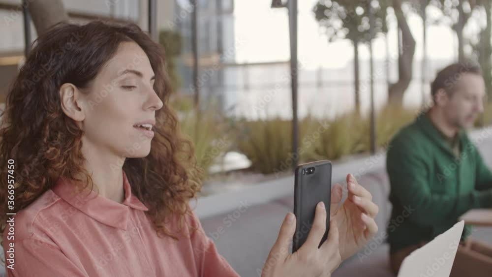 Tilt up of pretty curly-haired woman sitting at table in coworking space and making selfie on smartphone camera while middle-aged man working on laptop on background Tilt up of pretty curly-haired woman sitting at table in coworking space and making selfie on smartphone camera while middle-aged man working on laptop on background