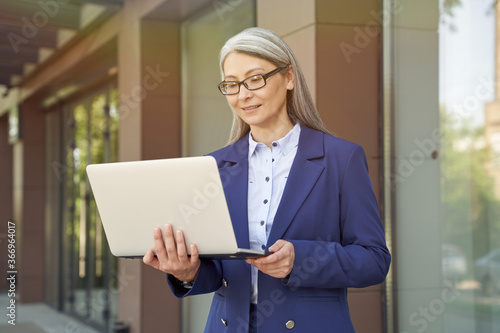 Waiting for a partner. Portrait of beautiful mature business woman wearing classic suit and eyeglasses looking at laptop screen while standing against office building outdoors