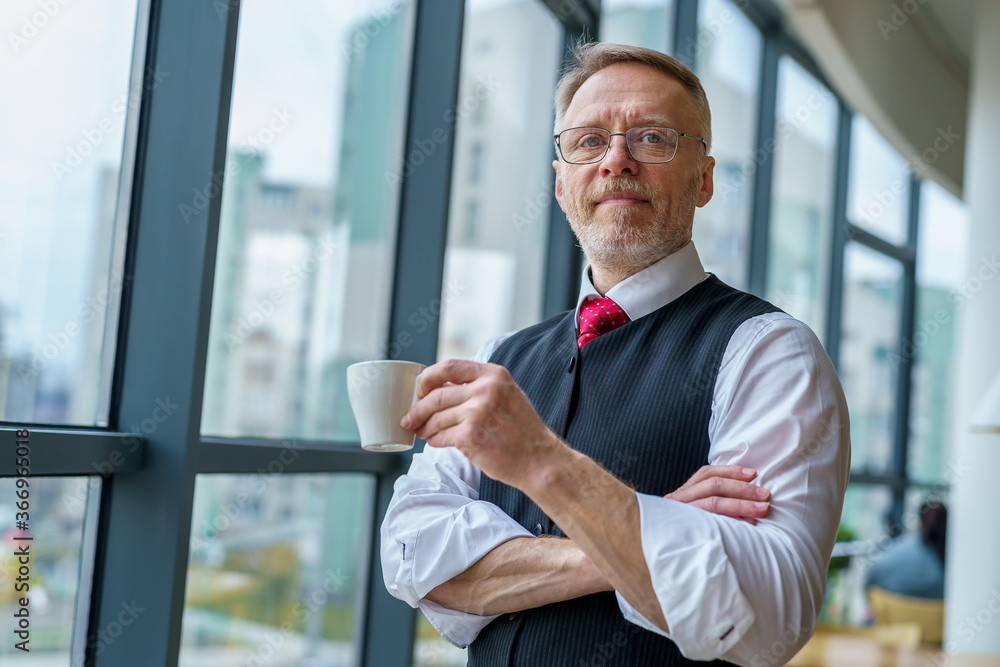 Senior businessman wears white shirt and drinking coffee near big window with city view . Photo from the side. Senior businessman wears white shirt and drinking coffee near big window with city view . Photo from the side.