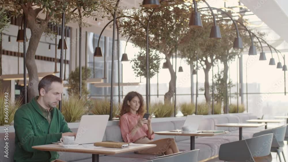 Panning of young curly-haired businesswoman sitting on sofa in cafe and using smartphone while middle-aged businessman sitting at table and working on laptop Panning of young curly-haired businesswoman sitting on sofa in cafe and using smartphone while middle-aged businessman sitting at table and working on laptop