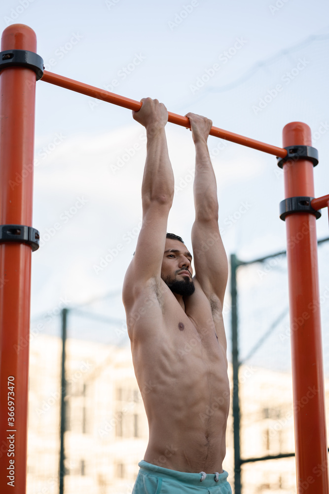 Fototapeta premium A muscular man does pull ups on a horizontal bar on a sports field