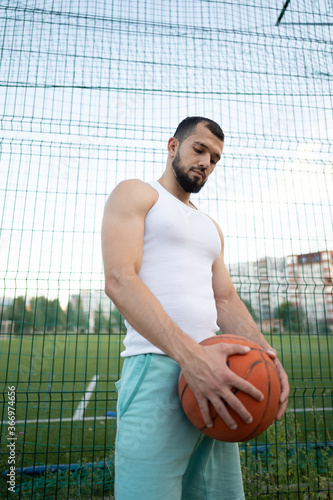 Man stands near a fence on the street, holding a basketball in his hand © Vladimir