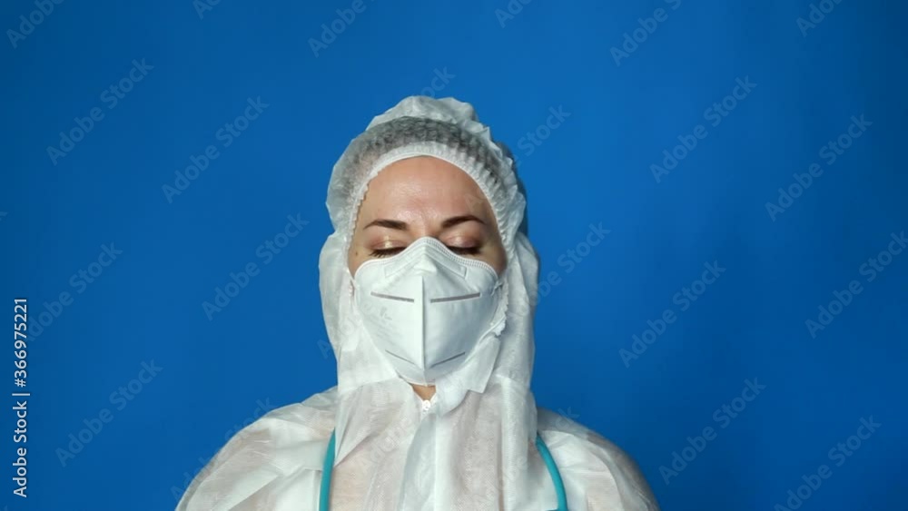 Tired female doctor in a protective suit and mask takes off his goggles and sighs on blue background. Doctors during COVID-19 concept. Tired female doctor in a protective suit and mask takes off his goggles and sighs on blue background. Doctors during COVID-19 concept.