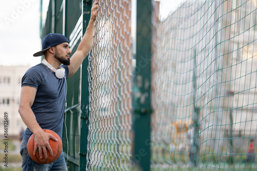 Man with a basketball looks out from behind a fence on the street during the day