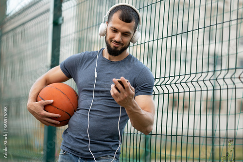 Man holds a basketball in his hand, looks at a smartphone and listens to music with headphones on the street
