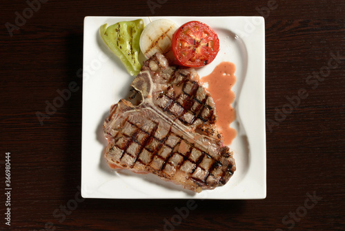 T-bone grilled beef steak with grilled vegetables of tomato, onion and bell pepper on a square plate on a wooden table. Top view. Photos for restaurant and cafe menus