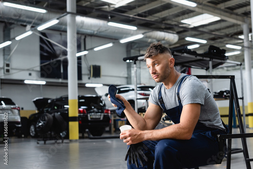 tired technician in overalls holding cap and disposable cup while sitting near cars in service station © LIGHTFIELD STUDIOS