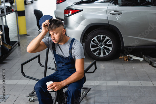 tired mechanic holding cap and paper cup while sitting near cars in garage