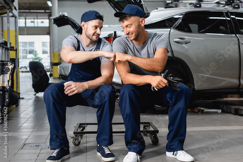 Smiling auto mechanic giving fist bump to colleague near autos at service station