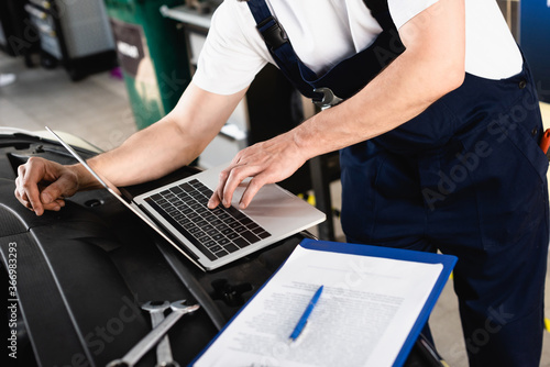 Cropped view of auto mechanic typing on laptop on hood near wrenches and clipboard at service station