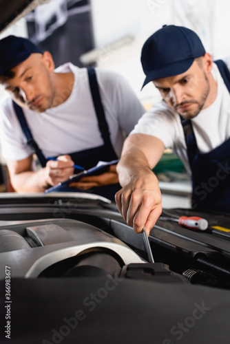 selective focus of mechanic in uniform repairing car near coworker with clipboard and pen