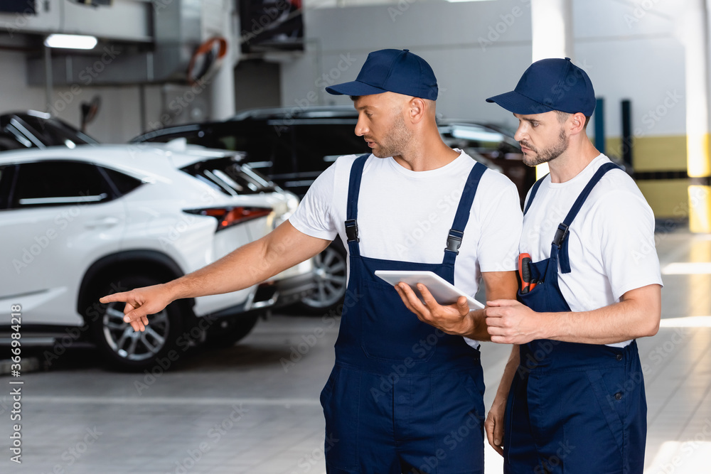 handsome mechanic in uniform holding digital tablet and pointing with finger at car near coworker in workshop handsome mechanic in uniform holding digital tablet and pointing with finger at car near coworker in workshop