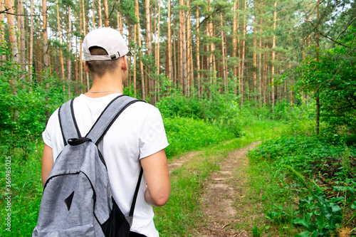 Back view of a male tourist in a white t-shirt cap with a gray backpack, walking along a path in the forest, turning his head, looking into the distance.