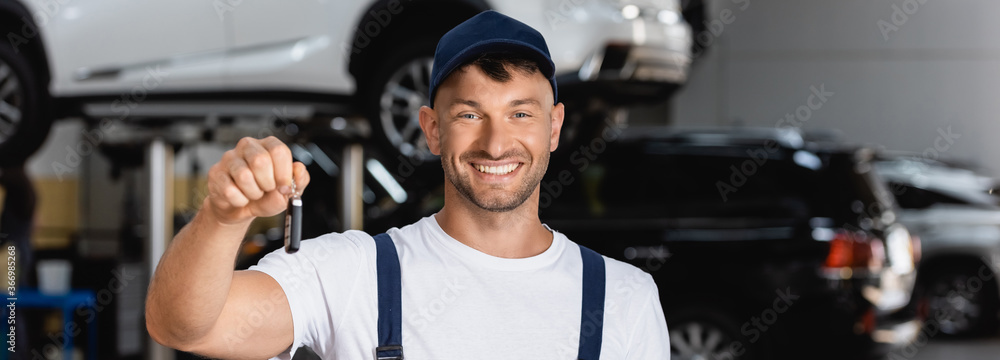 panoramic crop of cheerful mechanic in overalls and cap holding car key panoramic crop of cheerful mechanic in overalls and cap holding car key