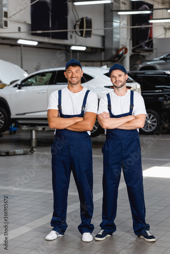 cheerful mechanics in uniform and caps standing with crossed arms in car service