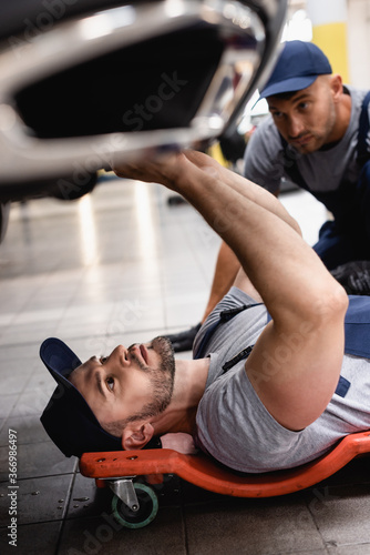 selective focus of mechanic lying and repairing auto near coworker in service center