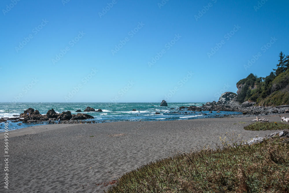 Oregon coastline with sand and grass foreground and surf and blue sky background.