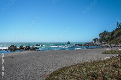 Oregon coastline with sand and grass foreground and surf and blue sky background.