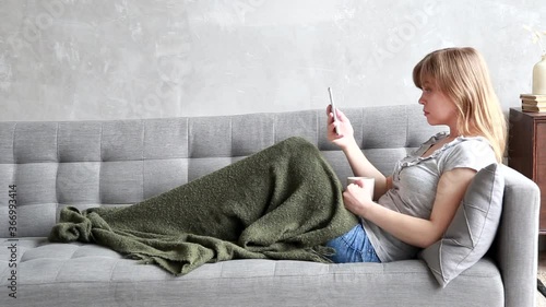 Blonde young woman sitting on the gray sofa and taking smartphone. The cozy staying at home. Sick girl lies under the covers. Covid-19 pandemic
