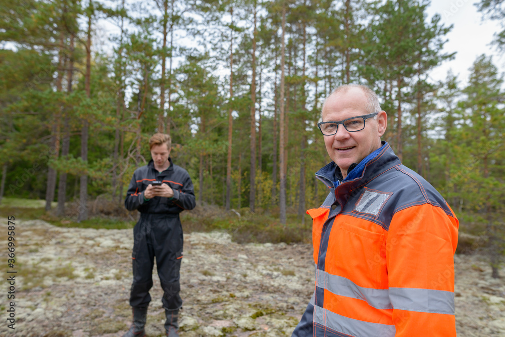 Happy mature man standing in the forest with young man using mobile phone in the background Happy mature man standing in the forest with young man using mobile phone in the background