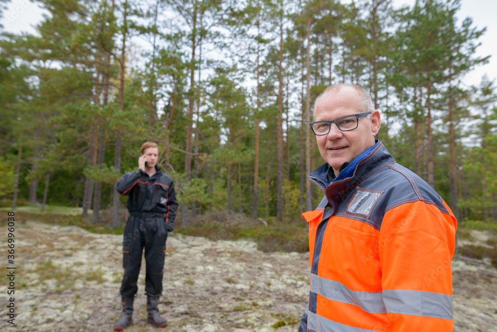 Happy mature man standing in the forest with young man talking on mobile phone in the background