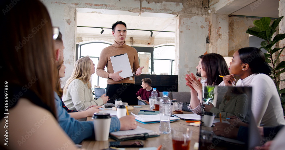 Obraz premium Happy young adult Asian business coach man giving a speech for multiethnic team at modern office workplace slow motion.