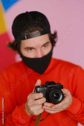 Male photographer. A young man of 25-30 years old in a black protective mask and in a red sweatshirt holds a mirrorless camera in his hands.  © Oleg Samoylov