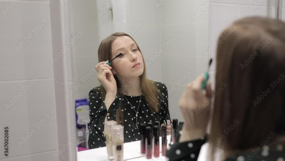 The girl is doing makeup and looking in the mirror in the bathroom.