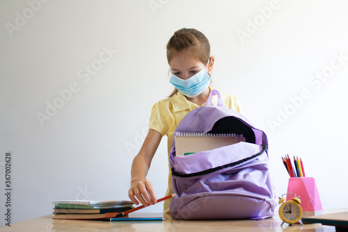 a schoolgirl wearing a face mask puts school supplies in a backpack