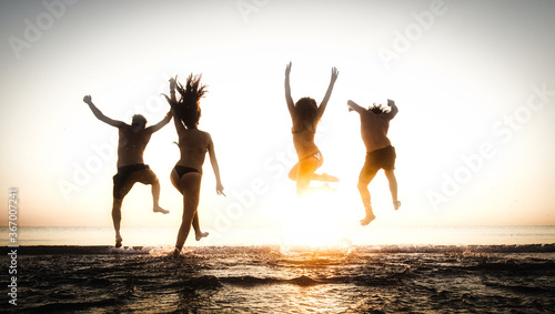 Happy group of people enjoying holidays jumping at the beach at sunset. Young joyful friends with arms up outdoor