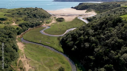 Aerial drone view flying down a beautiful green valley towards a huge sandy beach at low tide (Three Cliffs Bay, Swansea, Wales) photo