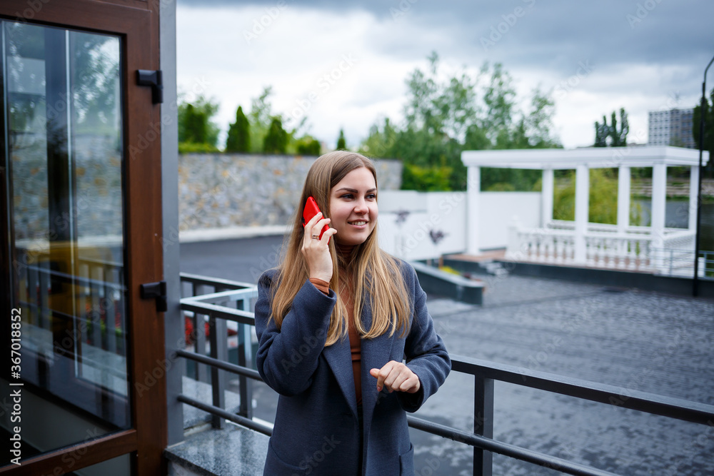 Fototapeta premium Young beautiful blonde woman in gray coat talking on the phone on a cold autumn day. Girl with phone on the terrace overlooking the river