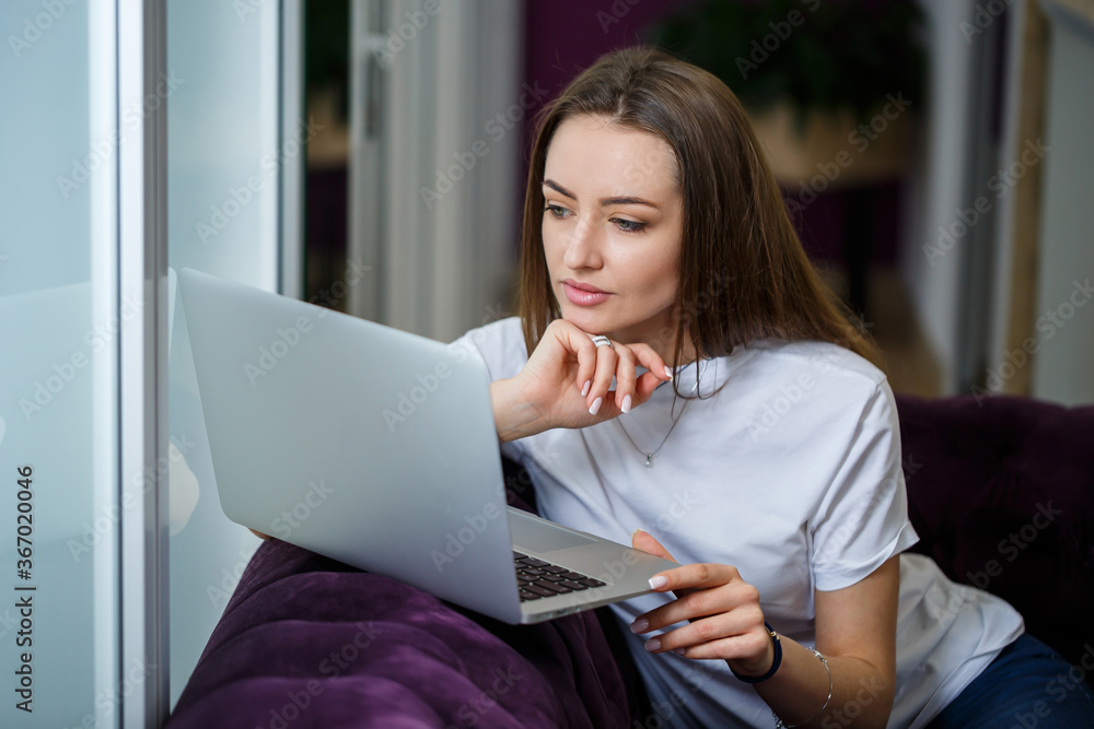 A young woman is sitting on the couch and working on a laptop remotely from work. Girl with a computer on her knees looking at the monitor A young woman is sitting on the couch and working on a laptop remotely from work. Girl with a computer on her knees looking at the monitor