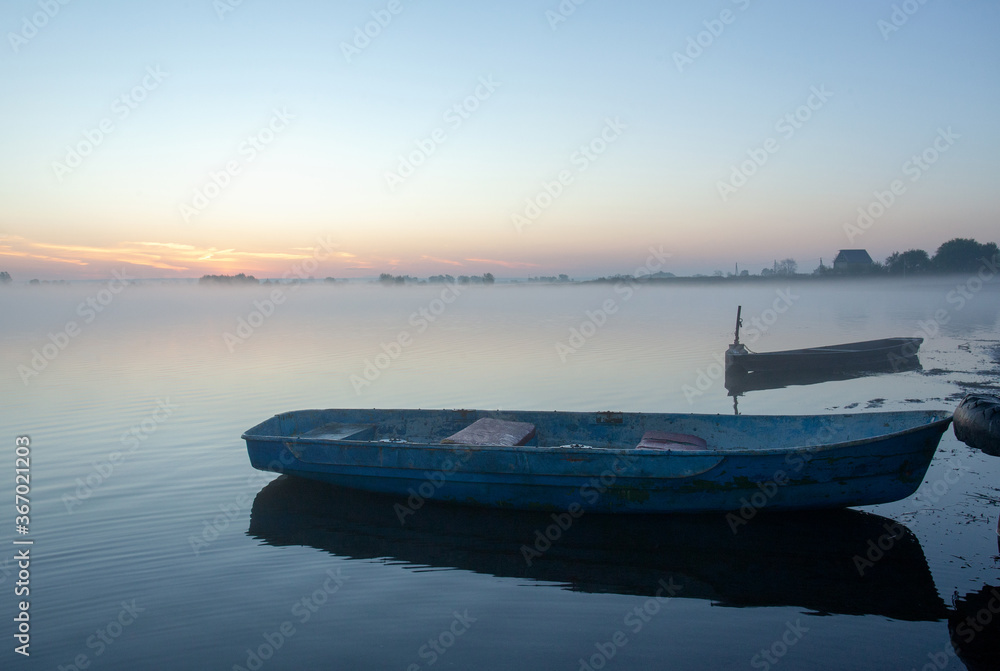 An early, foggy morning on the lake. Two boats at the shore. In the background outlines of houses. An early, foggy morning on the lake. Two boats at the shore. In the background outlines of houses.