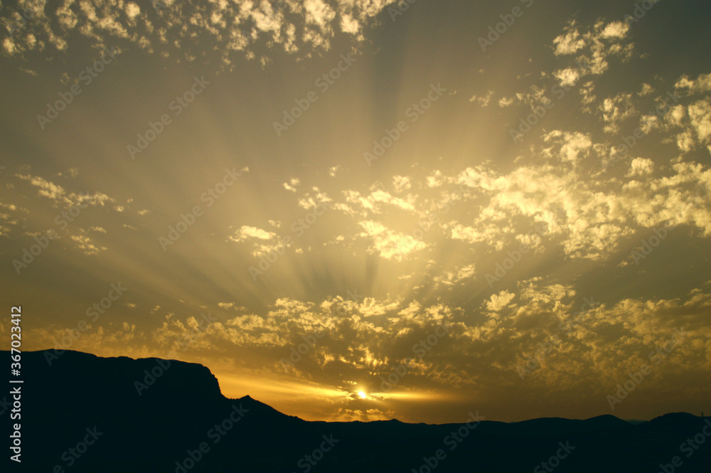 Sunset after a mountain with sun rays marked between the small clouds with yellow color