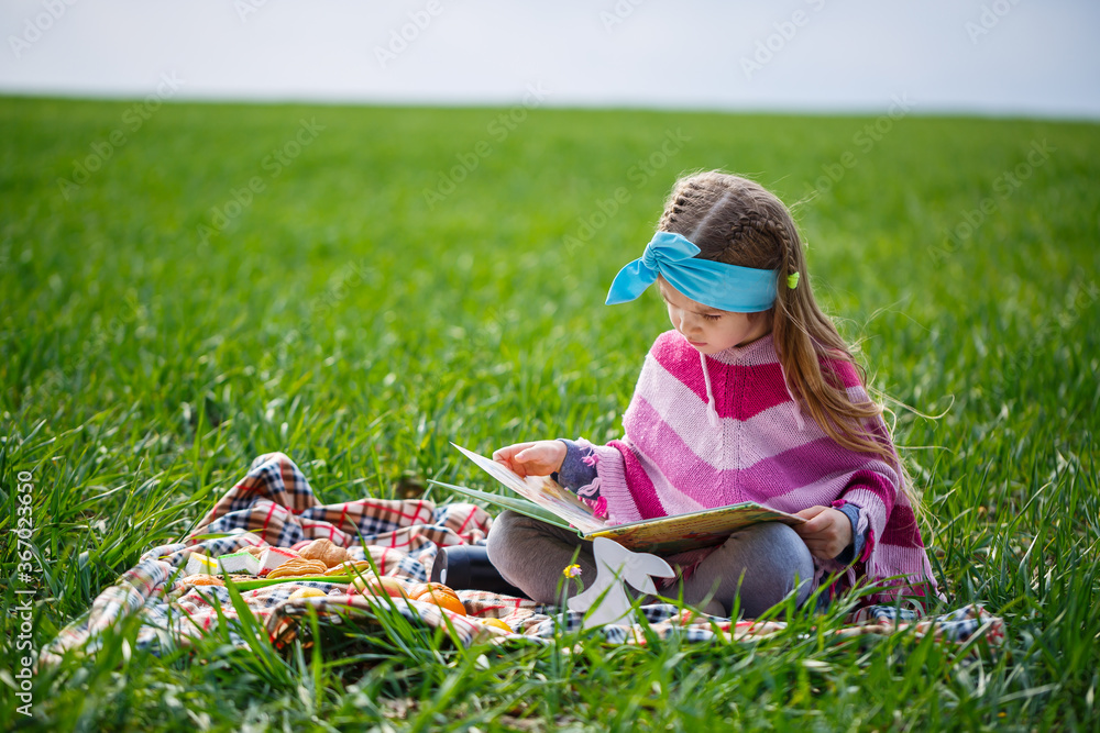 little girl child sits on a bedspread and reads a book with a fairy tale, green grass in the field, sunny spring weather, smile and joy of the child, blue sky with clouds