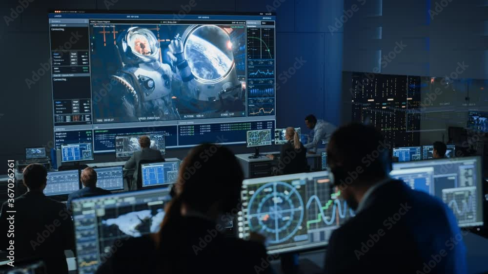 Group of People in Mission Control Center Establish Successful Video Connection on a Big Screen with an Astronaut on Board of a Space Station. Flight Control Scientists Sit in Front Computer Displays. Group of People in Mission Control Center Establish Successful Video Connection on a Big Screen with an Astronaut on Board of a Space Station. Flight Control Scientists Sit in Front Computer Displays.