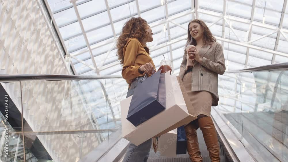Low angle view of two attractive Caucasian women wearing fashionable clothes standing in store with shopping bags and beverages in their hands and having chat Low angle view of two attractive Caucasian women wearing fashionable clothes standing in store with shopping bags and beverages in their hands and having chat