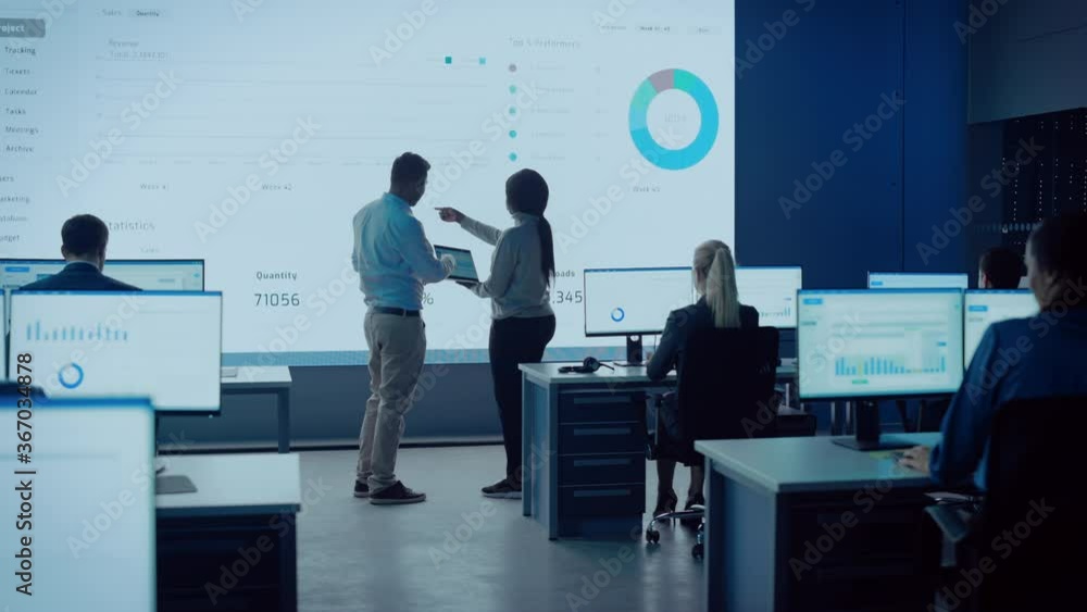 Two Traders Having a Meeting in a Modern Monitoring Office with Analytics Feed on a Big Digital Screen. Monitoring Room with Brokers and Finance Specialists Sit in Front of Computers. Colleagues Talk. Two Traders Having a Meeting in a Modern Monitoring Office with Analytics Feed on a Big Digital Screen. Monitoring Room with Brokers and Finance Specialists Sit in Front of Computers. Colleagues Talk.