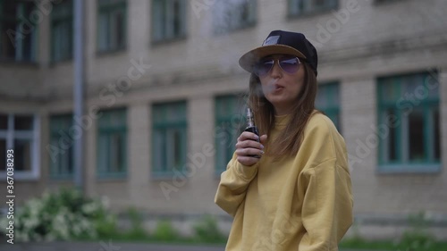 Pretty young hipster woman in black cap yellow hoodie vape ecig. Girl vape popular ecig gadget, vaping device.Happy brunette vaper girl with e-cig. Portrait of smoker female model with electronic photo