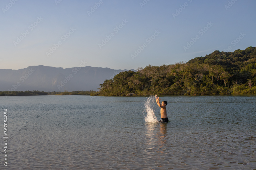 Menino brincando e se divertindo nas águas limpas do rio em linda tarde com por do sol