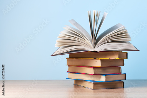 An open book lies on a stack of books that stands on a wooden table against a light blue wall, a place for text